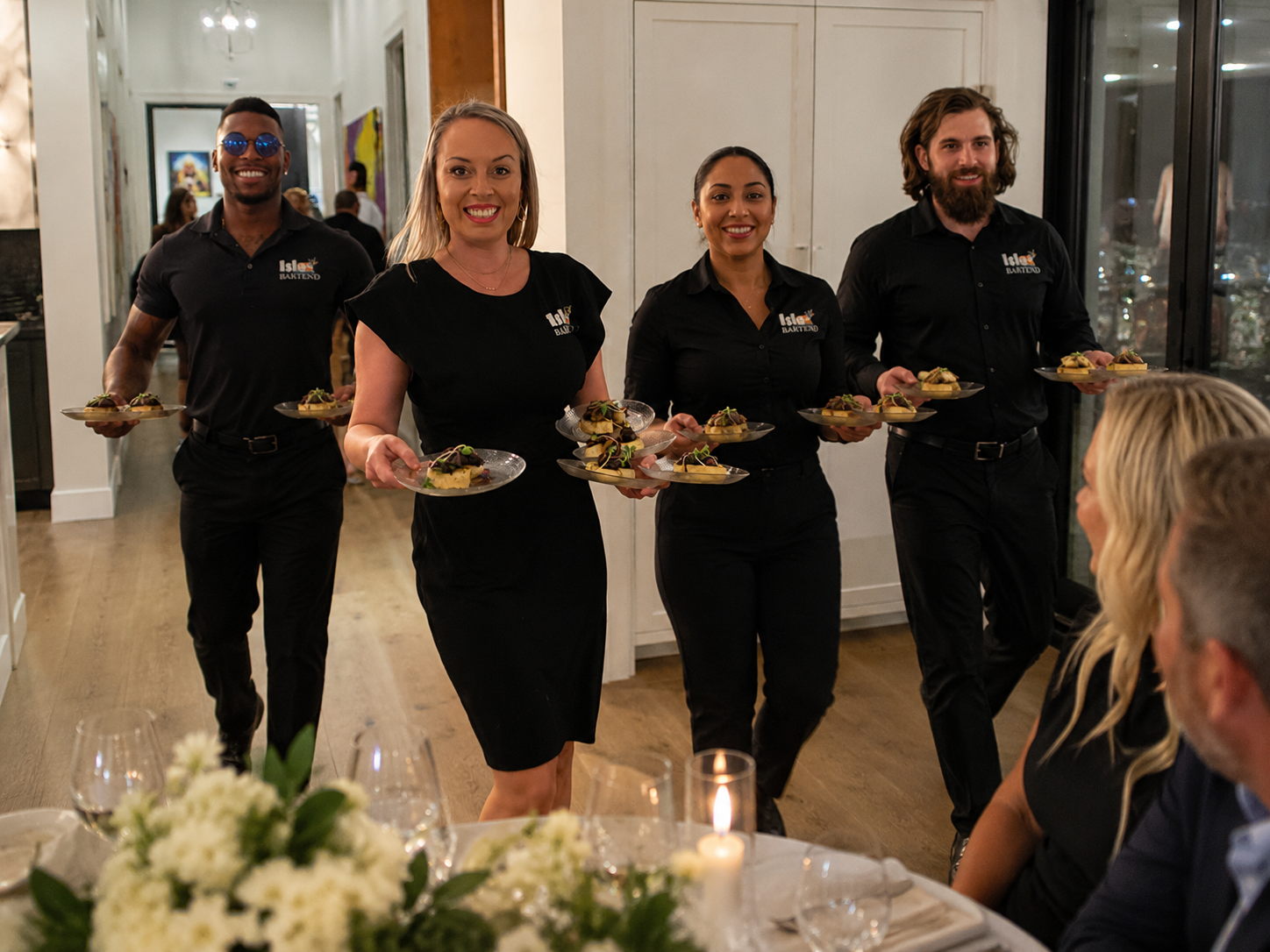 Four ISLE Bartend servers in black uniforms carrying plated courses to a candlelit table